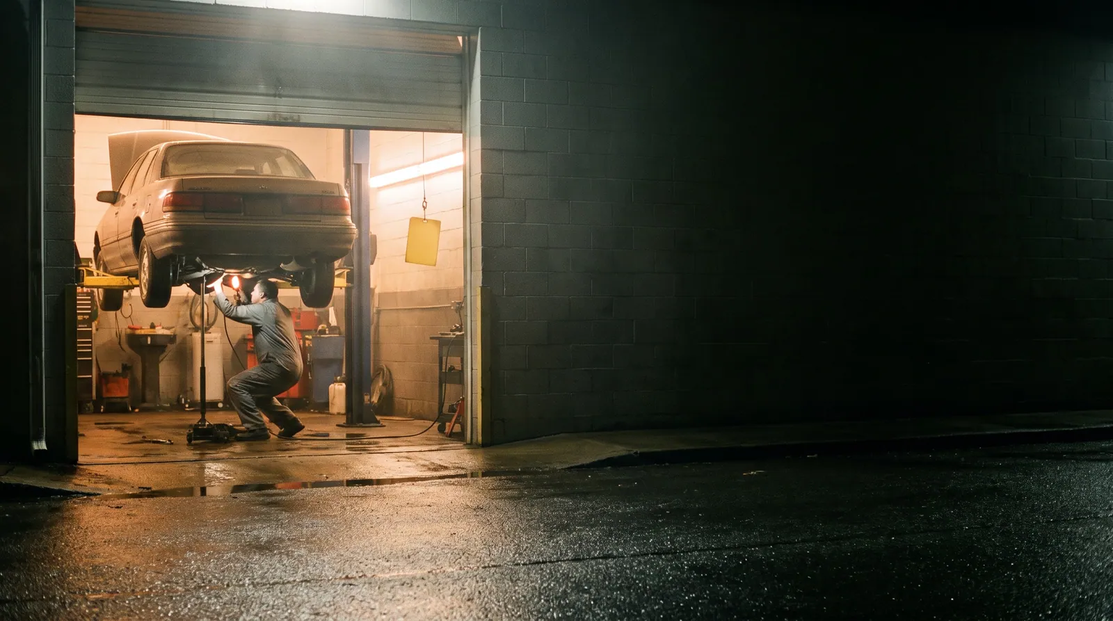 An independent repair shop at night after rain, one bay open and lit, a mechanic beneath a raised sedan