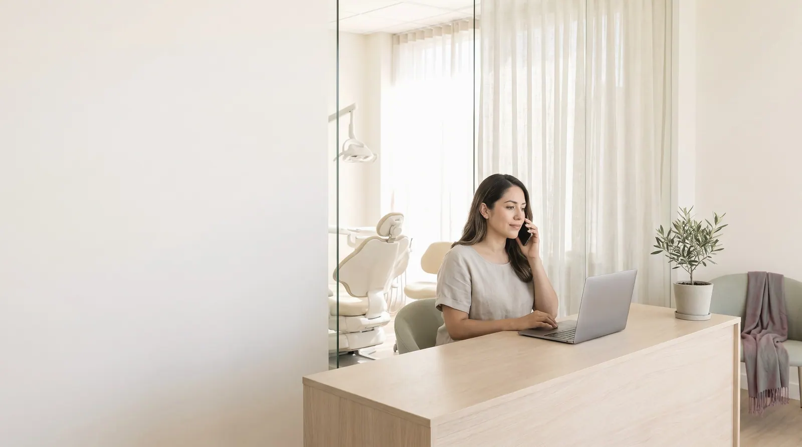 A front-desk coordinator on the phone at the pale oak reception desk of a bright dental studio