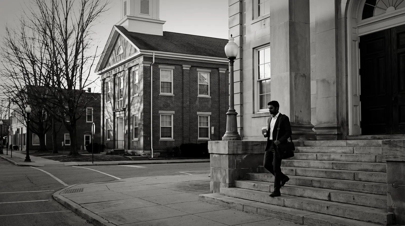 A young lawyer with a leather bag and a coffee walking down the steps of a small-town courthouse in the morning