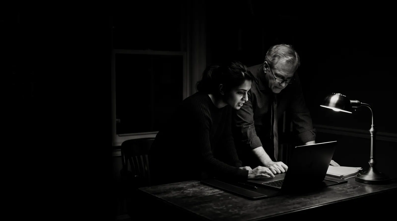 Late at night, an agency owner and a law firm partner lean over a laptop under a single desk lamp