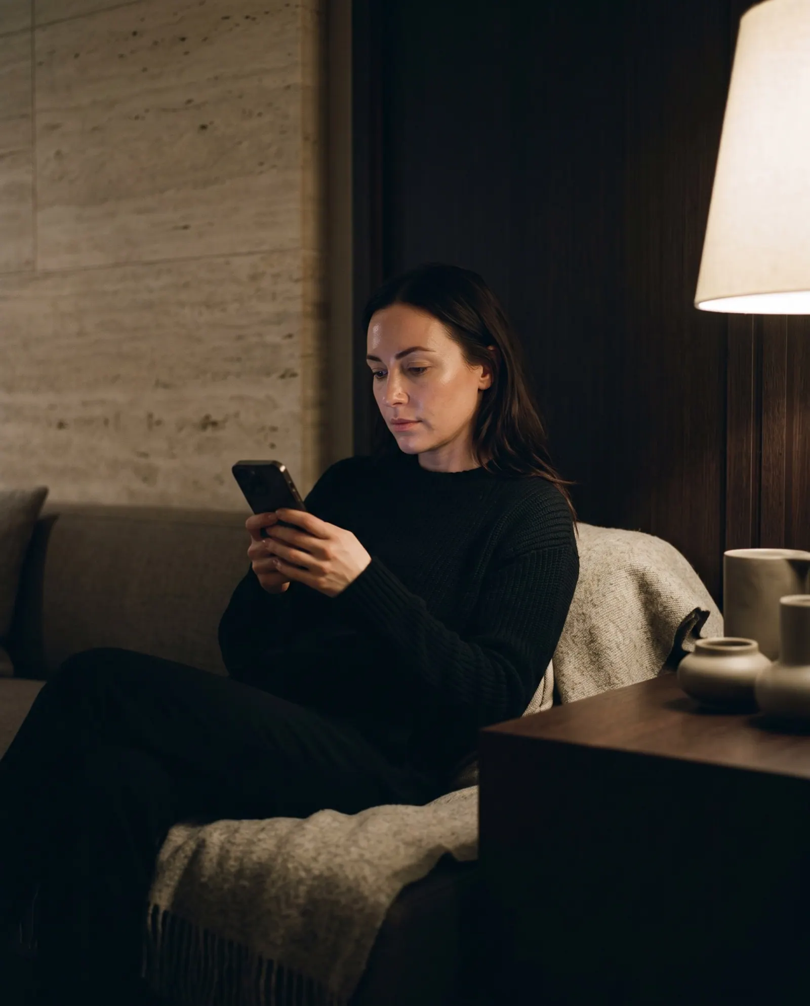 A woman on a sofa in lamplight reads a treatment page on her phone, deciding whether to book