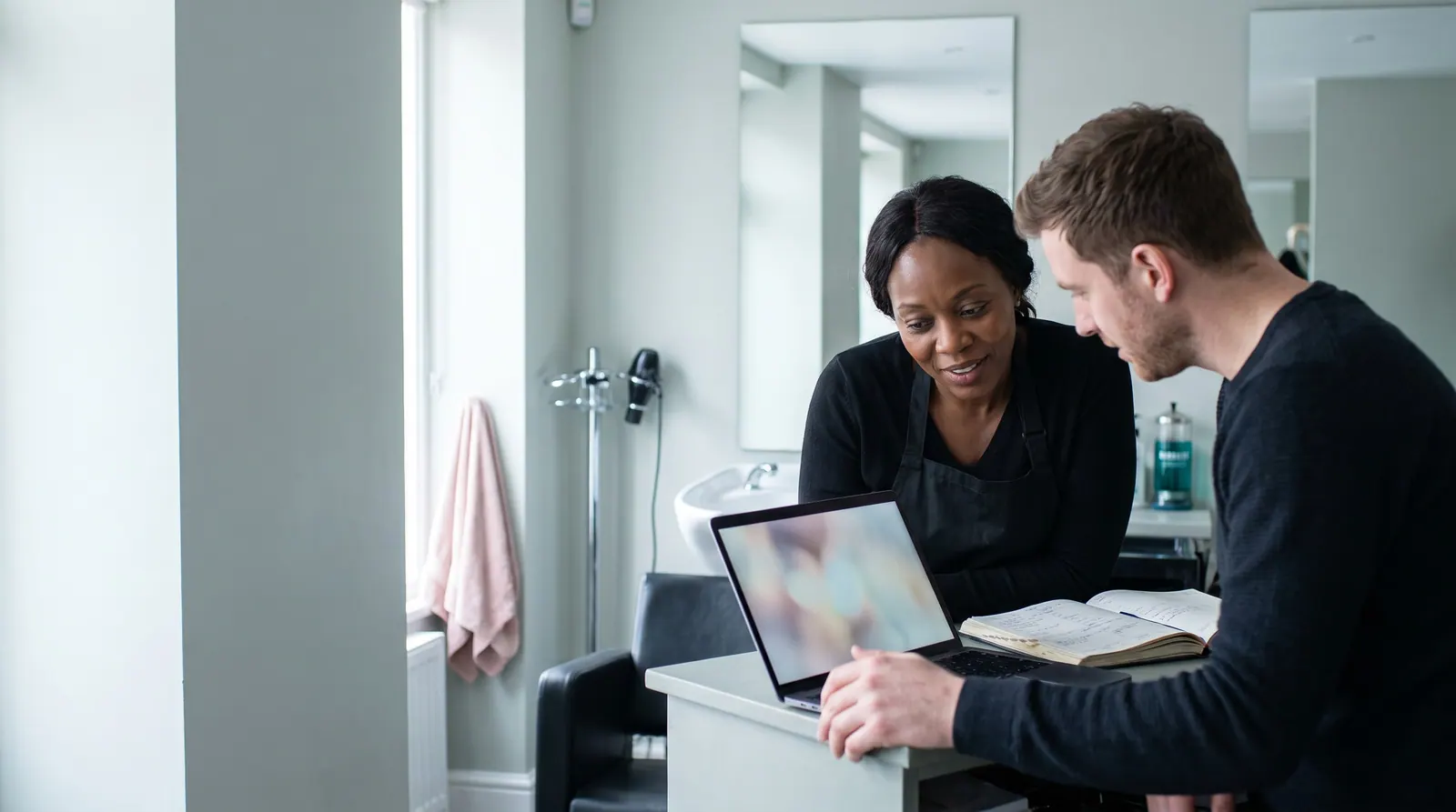 On a quiet morning before opening, an agency owner shows a laptop to a salon owner at the reception desk beside the appointment book